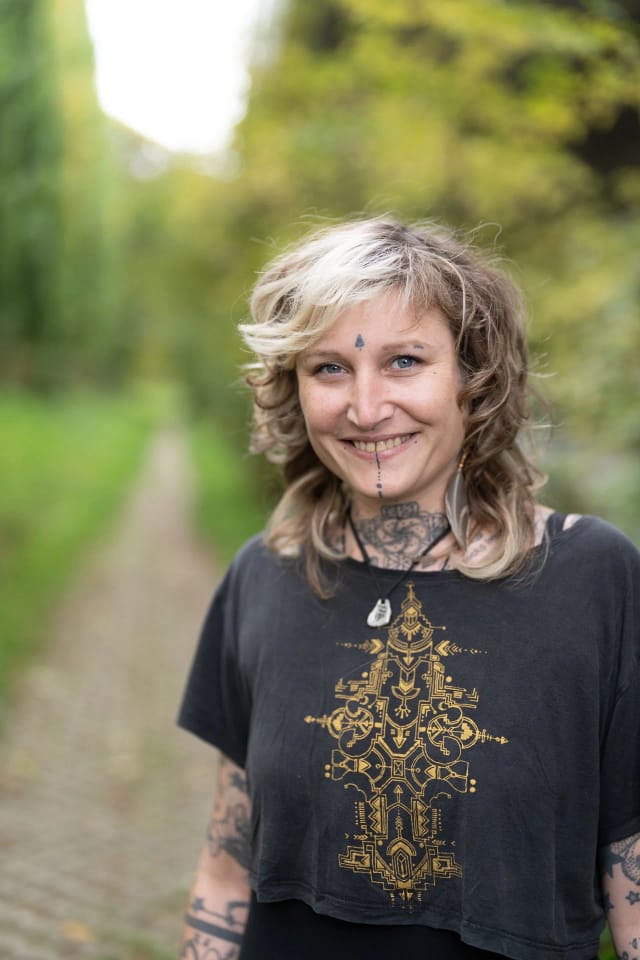 Lächelnde Frau im Freien mit Tätowierungen, schwarzen T-Shirt mit goldener Musterung, mehrfarbiger kurzer Haarfrisur, im Hintergrund grüner Waldpfad.